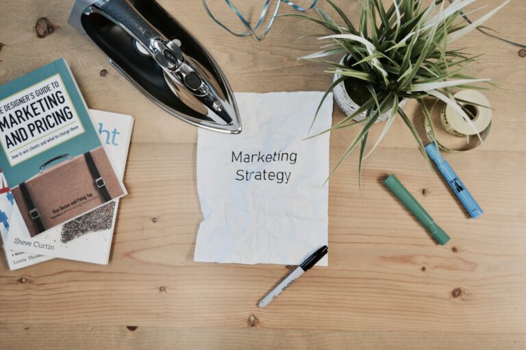 Paper labeled 'marketing strategy' surrounded by marketing books and assorted pens suggesting a workspace in preparation for marketing strategizing